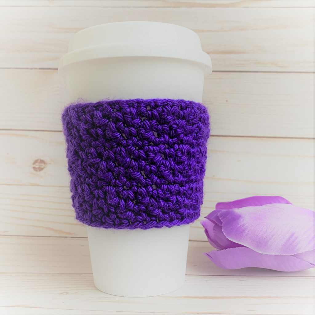 "Winter Hills Cozie" in purple on a reusable travel mug. Purple tulip in background